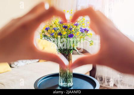 Frau zeigt Herz mit Fingern um blaue und gelbe Blumen. Kein Krieg in der Ukraine. Unabhängigkeitstag Symbol der Ukraine Stockfoto