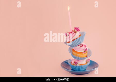 Drei Geburtstags-Beerenmuffins übereinander in blauen Wrappern mit einer Kerze darüber auf einer Untertasse auf rosa Hintergrund. Hochwertige Fotos für ca. Stockfoto