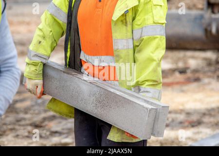 Nahaufnahme eines Bodenarbeiters in orangefarbener und gelber Hi-viz-Farbe, der während des Straßenbaus schwere Betonkanten auf der Baustelle trug Stockfoto