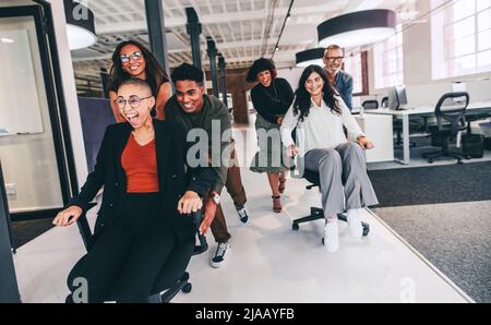 Viel Spaß im Büro. Fröhliche Gruppe von Kollegen, die sich in einem kreativen Büro auf Stühlen schieben. Ein Team von Geschäftsleuten, die Spaß haben und feiern Stockfoto