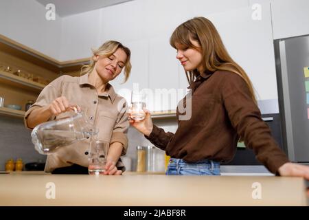 Mutter und Tochter Teenager trinken sauberes Wasser in der Küche und reden. Frau goss aus einem Krug Wasser in ein Glas Stockfoto