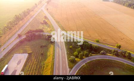 Flugdrohne Flug über Auto Austausch zwischen landwirtschaftlichen Weizenfeldern am frühen Sommermorgen. Landstraße, Autos, Felder, Bäume, Morgennebel, Horizont Sommer Sonnenaufgang. Landschaftlich reizvoll Stockfoto