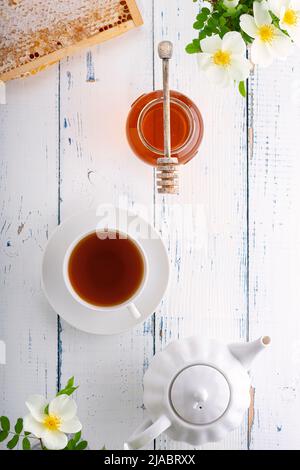 Honig im Glas, ein Holzlöffel für Honig, Waben, Tee in einer weißen Tasse und Hagebutten auf einem Holztisch. Draufsicht. Stockfoto