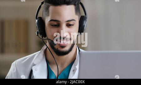Professionelle männliche arabische hispanische junge Ärztin im medizinischen Mantel tragen Headset mit Mikrofon Machen Sie Telefonkonferenz auf Laptop-Nutzung Entfernung Stockfoto