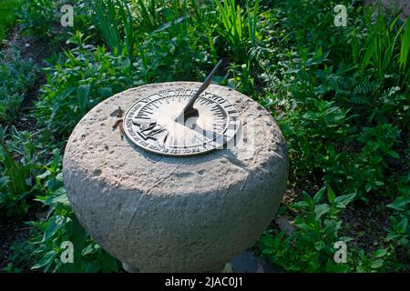 Metallische Sonnenuhr in Stein auf einem Bett verschiedener grüner Pflanzen und Gräser an einem sonnigen Tag Stockfoto