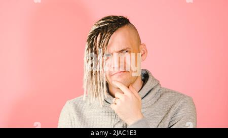 Junger nachdenklicher, hübscher Mann mit blonden Dreadlocks auf rosa Hintergrund. Nachdenklicher Kerl mit Zweifel Blick klopft sein Kinn mit dem Finger Stockfoto