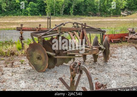 Ein rostiger alter Zug hinter einer Traktorfräse, die im Freien auf dem Feld steht und von anderen landwirtschaftlichen Werkzeugen umgeben ist Stockfoto