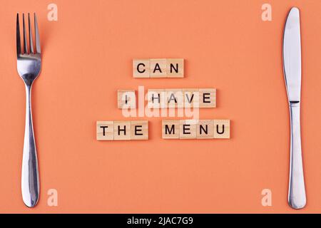 Gabel, Messer und Text kann ich das Menü auf Holzwürfel schreiben lassen. Flache Lay-Komposition auf orangefarbenem Hintergrund. Stockfoto