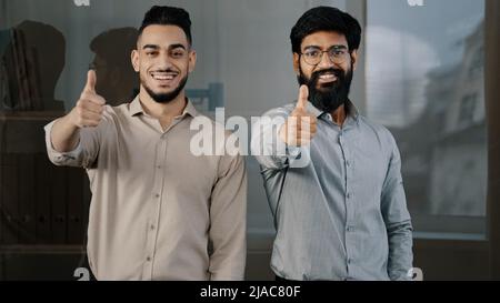 Glückliche selbstbewusste Männer junge Berufskollegen Mitarbeiter Partner arabische hispanische Geschäftsleute, die auf die Kamera schauen und dabei Daumen nach oben zeigen Stockfoto