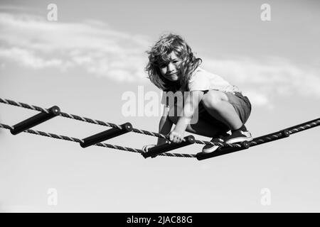 Netter Junge klettert die Leiter auf dem Spielplatz hoch. Kind klettert die Leiter gegen den blauen Himmel hinauf. Schöner lächelnder netter Junge auf einem Spielplatz. Kinder Stockfoto