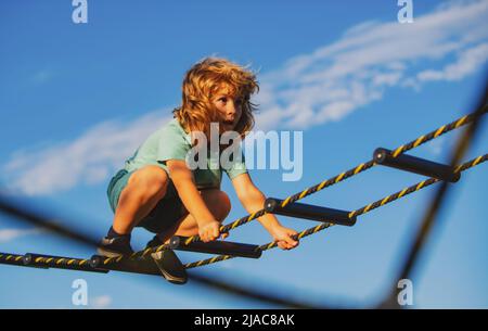 Netter Junge klettert die Leiter auf dem Spielplatz hoch. Kind klettert die Leiter gegen den blauen Himmel hinauf. Kleiner Junge trainieren auf dem Spielplatz. Stockfoto