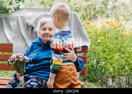 Enkeljunge, der der Großmutter eine Blume gibt. Enkel und Großmutter verbringen Zeit miteinander. Oma mit Enkeln genießen Zeit zusammen im Freien Stockfoto