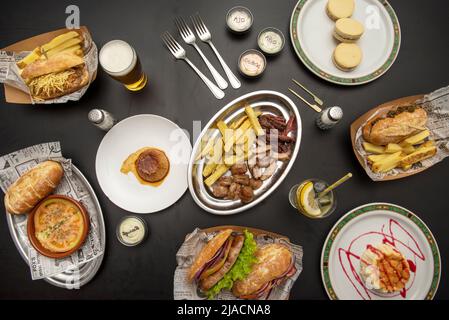 Auswahl an Tellern mit Sandwiches, pommes frites, Roastbeef in Scheiben, Panna Cotta, Eierpudding und Käsekuchen auf schwarzem Tisch Stockfoto