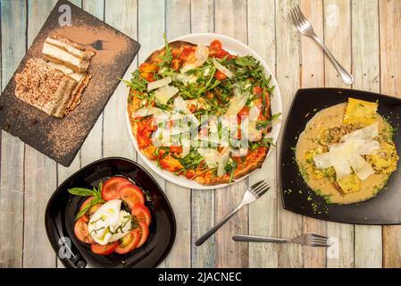 Set aus italienischen Gerichten mit neapolitanischer Pizza mit viel Rucola, Burrata-Salat mit Tomaten und Olivenöl, Ravioli mit Käse und Sauce und Tiramisu Stockfoto