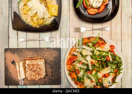 Italienische Gerichte mit neapolitanischer Pizza mit viel Rucola, Burrata-Salat mit Tomaten und Olivenöl, Ravioli mit Käse und Sauce und Tiramisu Stockfoto