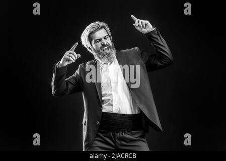 Fröhlicher, junger Geschäftsmann in schwarzem Anzug, der Musik mit Kopfhörern hört und auf grauem Hintergrund tanzt. Stockfoto