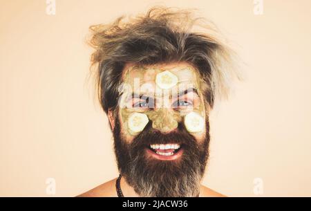 Happy man mit lustigen Gesichtsmaske mit Gurke Spaß haben. Verfahren zum Auftragen der Maske aus Ton auf das Gesicht des lustigen Mannes. Spa im Haus. Stockfoto