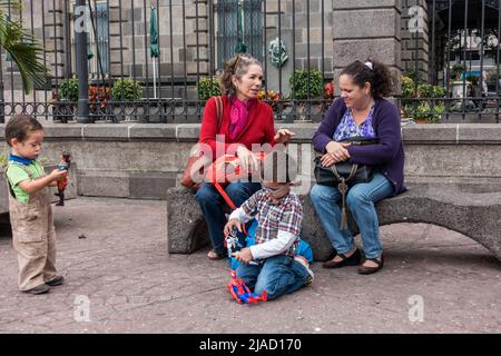 Zwei Frauen unterhalten sich auf einer Parkbank, während zwei Kinder in San José, Costa Rica, mit ihren Spielsachen spielen. Stockfoto