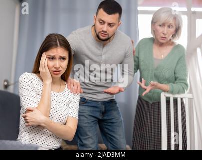 Mann, der auf dem Sofa sitzt, seine Frau und seine Schwiegermutter streiten sich mit ihm Stockfoto