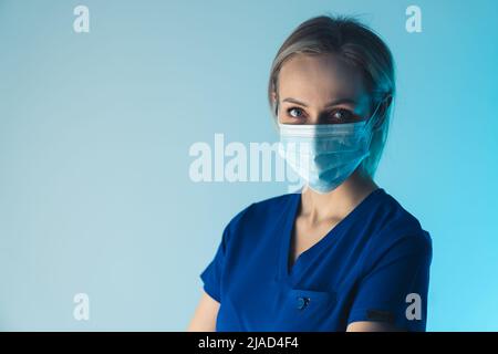 Attraktive konzentrierte europäische Hausärztin in einer OP-Maske und einem blauen Medizin-T-Shirt, das direkt auf die Kamera schaut. Nahaufnahme im Studio. Hochwertige Fotos Stockfoto