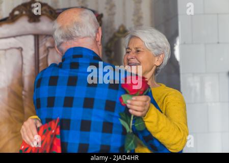Glückliche und dankbare Großmutter mit Geschenken umarmt ihren Mann mittlere Nahaufnahme Wohnzimmer Dienstalter und Urlaubskonzept. Hochwertige Fotos Stockfoto