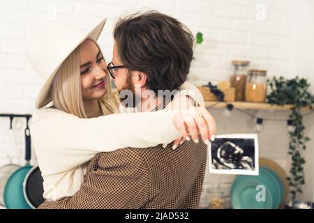 Innenaufnahme eines hübschen, tausendjährigen kaukasischen Freundes und einer Freundin mit einem Ultraschallbild ihres zukünftigen Neugeborenen, das in einer Küche steht und sich umarmt. Hochwertige Fotos Stockfoto