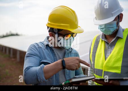 Zwei Ingenieure, die Gesichtsmasken tragen und ein digitales Tablet in einer Solarstation in Thailand betrachten Stockfoto