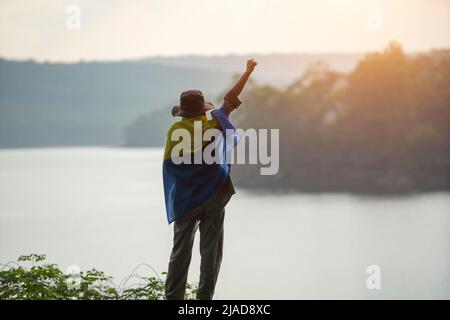 Rückansicht einer Frau, die mit der Hand in der Luft in eine ukrainische Flagge gehüllt ist, Thailand Stockfoto