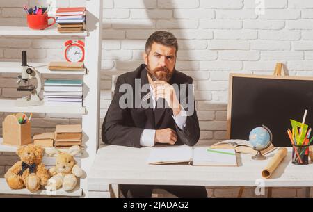 Porträt eines männlichen Lehrers aus der Vorschule oder Grundschule, der am Schreibtisch in der Klasse arbeitet. Reifer Mann sitzt entspannt in einem Klassenzimmer Blick auf die Stockfoto