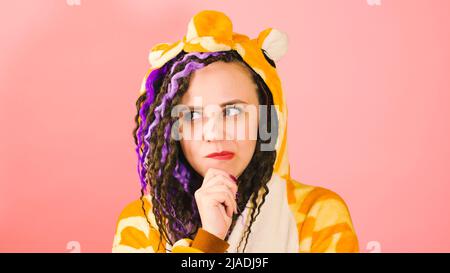 Junge, nachdenkliche Frau in Kigurumi kratzt sich am Kinn und schaut sich auf rosa Hintergrund um. Niedliche lebendige Frau mit Dreadlocks im Schlafanzug mit reflektierenden Th Stockfoto