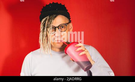 Junger hübscher stilvoller Mann in Gläsern, schwarze Kappe trinkt erfrischendes Getränk auf rotem Hintergrund. Hipster mit blonden Dreadlocks trinken Softdrink Th Stockfoto