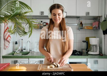 Frau mit Kopfhörern schneidet Banane für gesunden Salat oder Smoothie Stockfoto