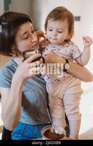 Affektive Mutter hilft ihrer kleinen Tochter, ein Glas Milch zu trinken, das sie an ihren Armen hält. Schöne junge Frau. Babypflege. Stockfoto