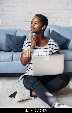 afroamerikanische Frau, die zuhause mit einem Computer-Laptop aus der Ferne studiert und dabei über Fragen nachdenkt, auf dem Boden sitzt. Lockiges schwarzes Haar Stockfoto