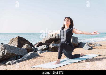 Voller Inhalt Flexible weibliche Steh in Anjaneyasana mit ausgestreckten Armen auf Matte am Sandstrand während des Yoga-Trainings im Sommer Stockfoto