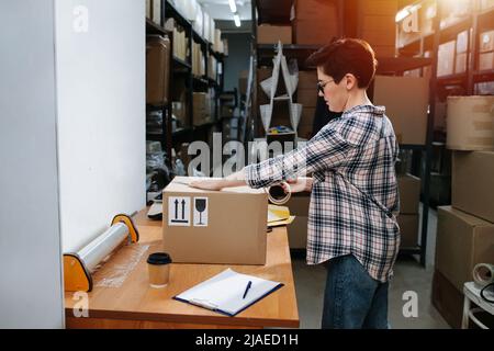 Seitenansicht einer kurzhaarigen Lagerarbeiterin, die einen Karton mit einem Messlöffel Klebeband auf einen Tisch packte. Sie trägt eine Brille und ein kariertes Hemd. Stockfoto