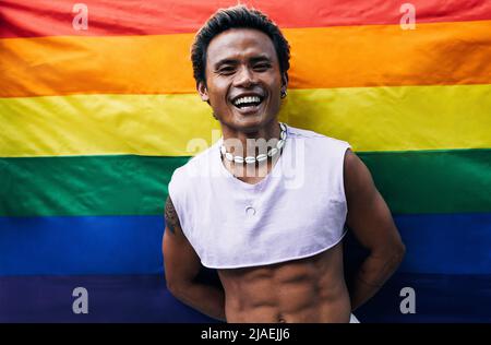 Fröhlicher Kerl, der auf der LGBT-Flagge posiert. Lachender Mann, der gegen die Regenbogenfahne steht. Stockfoto