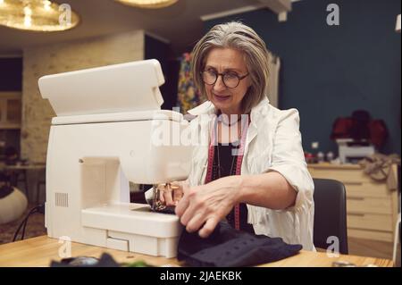 Stilvolle und modische Kleidung Designerin ältere Frau näht an einer Nähmaschine. Nähen, Designarbeiten, Schneideratelier, Schneider, Designer-Kleidung, manu Stockfoto