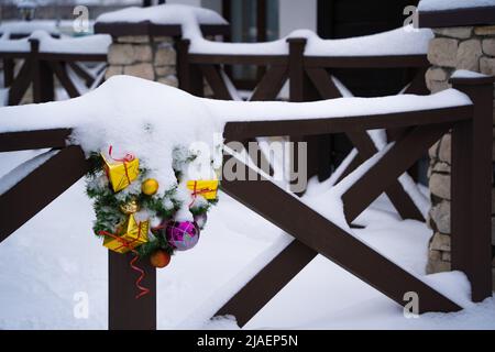 Blick auf den Winter - schneebedeckter weihnachtskranz, der auf einem hölzernen Geländer auf der Veranda mit Steinsäulen hängt Stockfoto