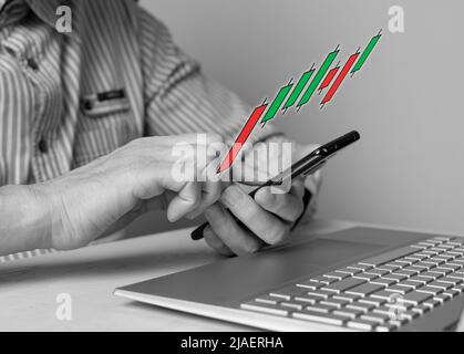 Mann mit Telefon in den Händen, der Candlestick-Graph verwendet, um die Kursbewegung zu verfolgen. Börsenanalyse, Investment, Handel, Finanzierungskonzept. Mann sitzt am Tisch mit Laptop. Schwarzweiß. Foto Stockfoto