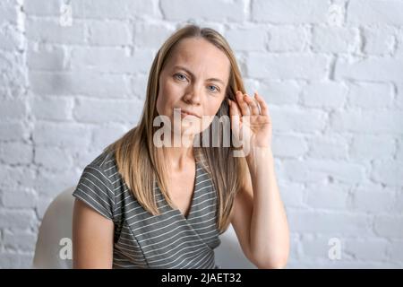 Porträt mit Schuss eine fröhliche blonde Frau mittleren Alters, die lächelt, wenn man die Kamera anschaut, fühlt sich in einer glücklichen Pose an, blauäugig in einem grauen Kleid an einer weißen Ziegelwand, Nahaufnahme Stockfoto