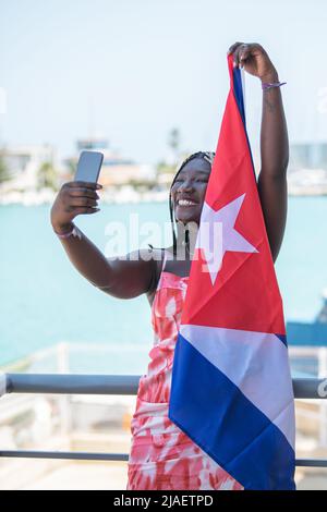 Frau mit kubanischer Flagge nimmt ein Selfie mit dem Meer und der Stadt dahinter Stockfoto