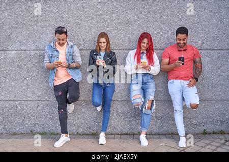 Eine Gruppe von multiethnischen Freunden, die ihr Mobiltelefon benutzen, während sie sich an einer Wand auf der Straße lehnen. Technologie, Freundschaft und urbanes Lifestyle-Konzept. Stockfoto