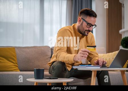 Porträt eines jungen, erwachsenen Unternehmers, der online am Laptop arbeitet und in seinem Wohnzimmer mit Kreditkarte sitzt. Geschäftsmann beim Stöbern in der inte Stockfoto