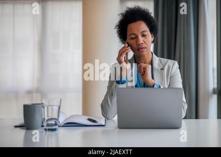 Geschäftsfrau im Gespräch auf dem Smartphone. Die Managerin arbeitet am Laptop, während sie am Schreibtisch sitzt, sie ist in ihren Formulalen im Büro der Firma. Stockfoto