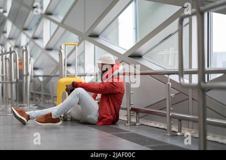 Aweary junger afroamerikanischer Mann sitzt mit dem Smartphone nach der Ankündigung der Flugstornierung Stockfoto