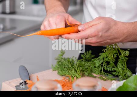 Mann bereitet vegetarisches Essen zu Essen Peeling Karotte auf Schneidebrett. Hausgemachte Mahlzeit Stockfoto