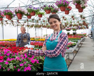 Junge lächelnde weibliche Floristin mit Gartenschürze im Gewächshaus, die Blumen im Topf zeigt, während der Mann im Hintergrund steht. Stockfoto