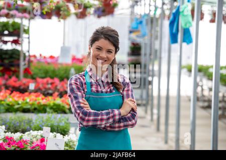Porträt einer hübschen Frau mit Schürze, die im Frühjahr mit gekreuzten Armen in der Baumschule mit verschiedenen Blumen im Hintergrund steht Stockfoto