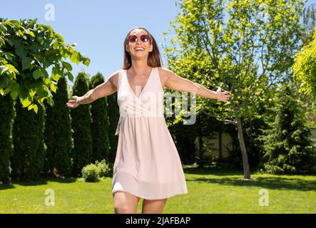 Lächelnde Frau mit Sonnenbrille genießt den sonnigen Tag und fühlt sich frei, während sie mit den Armen zum Himmel tanzt. Stockfoto